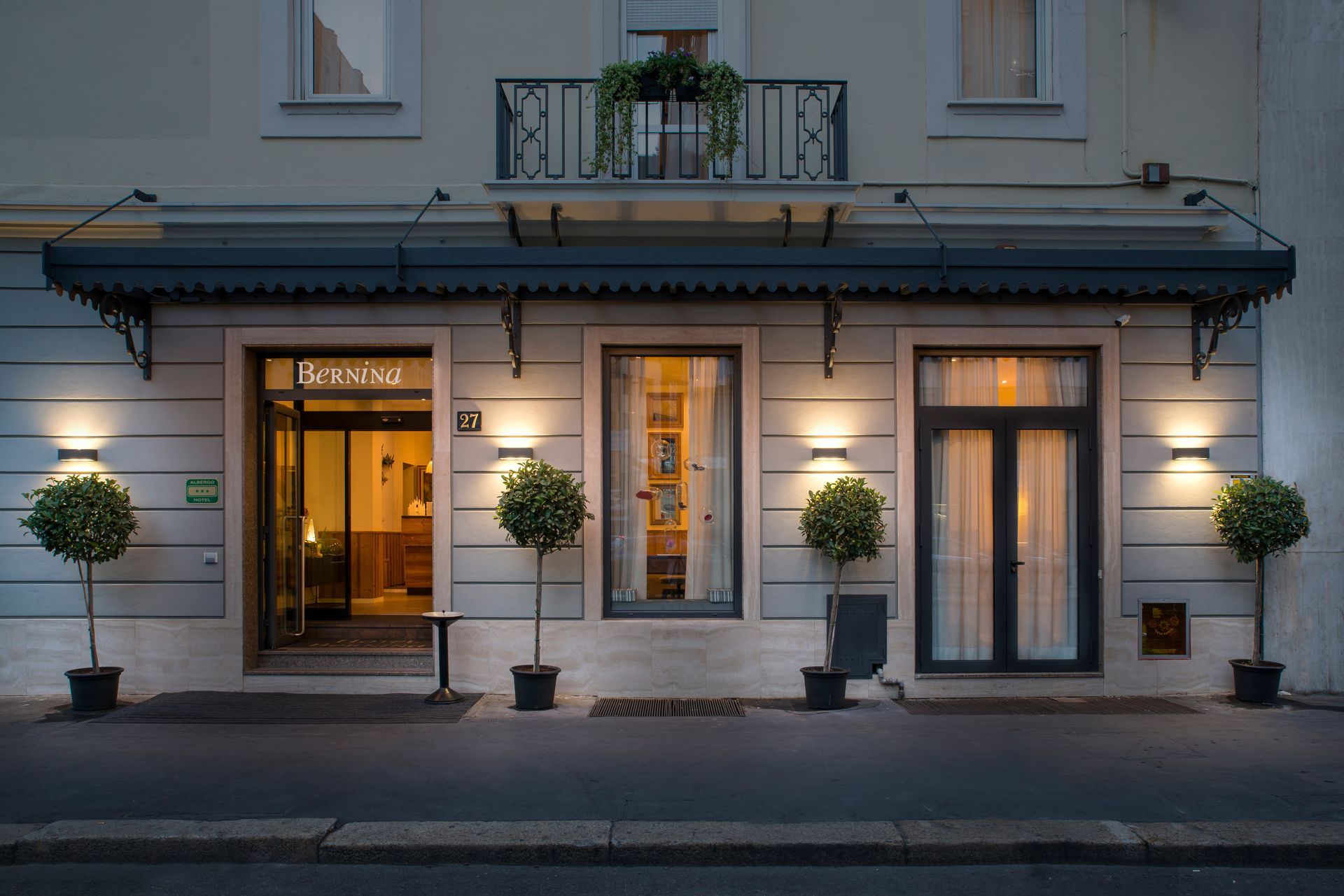 Exterior de un edificio al anochecer con entradas iluminadas. Árboles en macetas flanquean las puertas y ventanas.