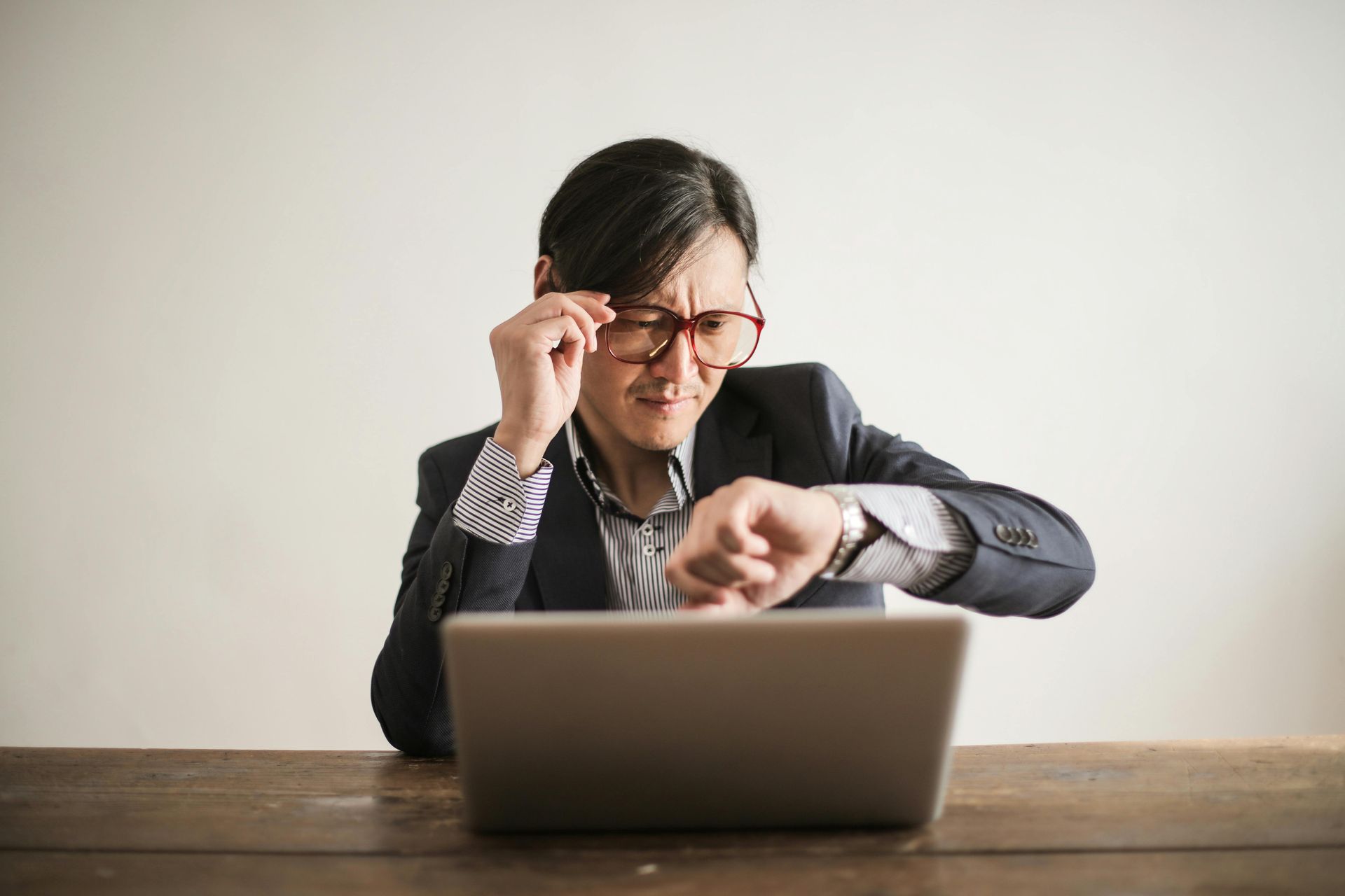 Man in glasses checks watch, looking at laptop, seated at a wooden desk.