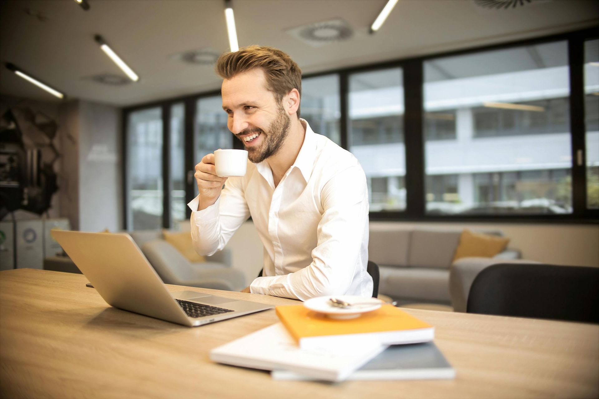Man in white shirt smiling, sipping from a cup, working on laptop at desk in office.