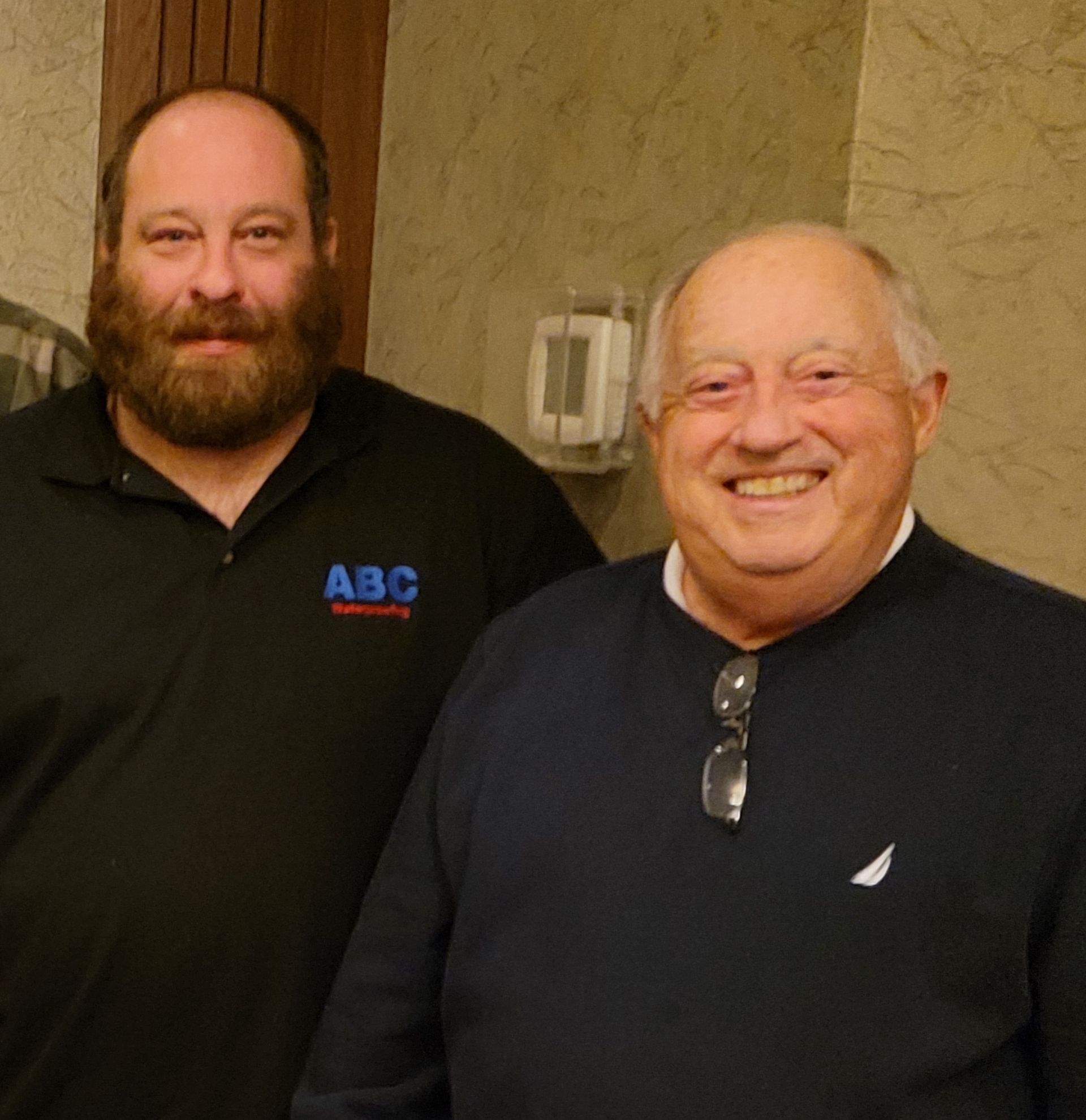 Two men smiling, one with a beard and a black shirt, the other in a blue sweater, indoors.