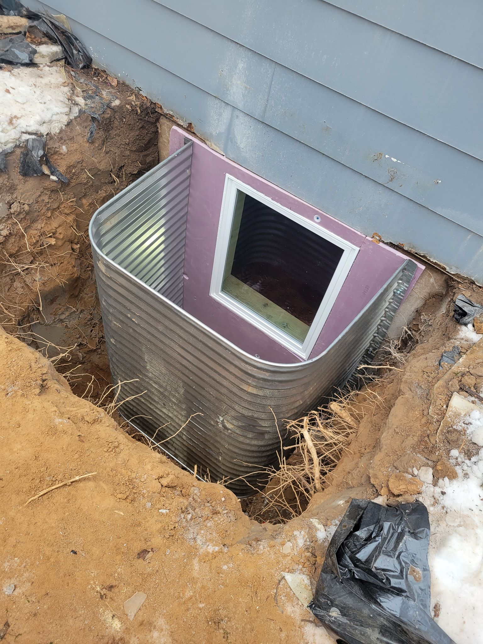 Metal window well in excavated ground next to a gray house with a window, pink insulation.