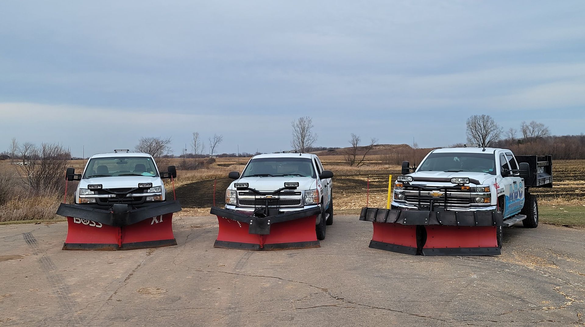 Three white pickup trucks with snowplows, parked outdoors.