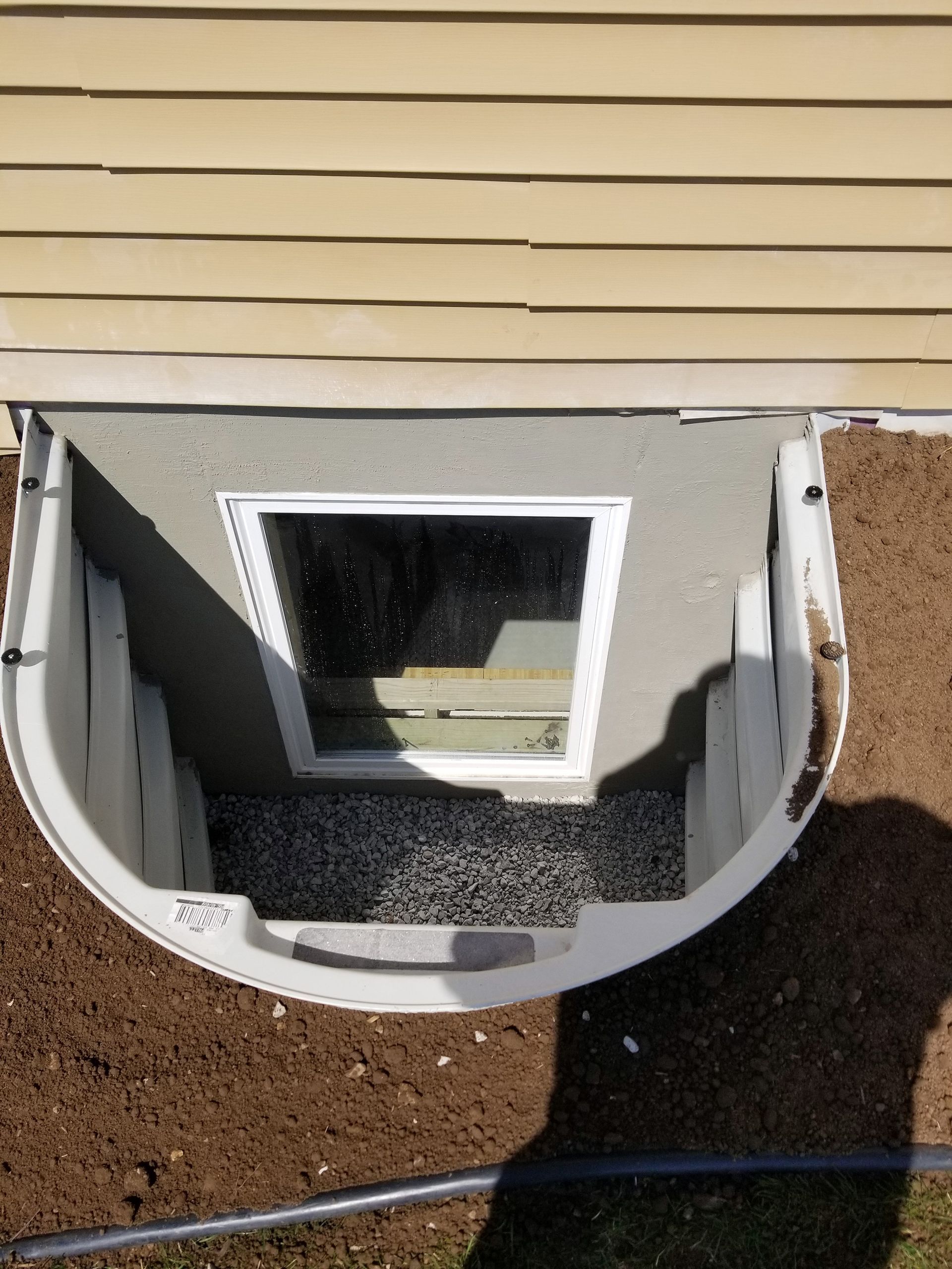 White window well with window and steps; beige siding above, brown soil around.
