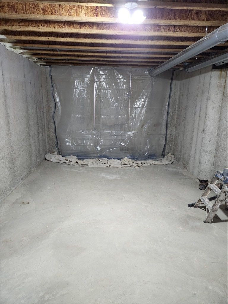 Concrete basement with plastic sheeting on one wall, under exposed wooden beams and a light fixture.