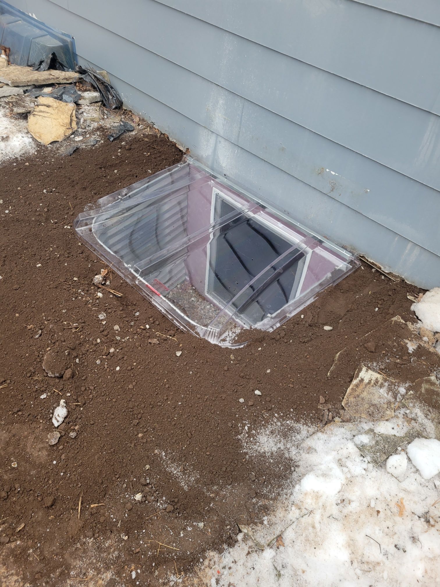 Window well with clear plastic cover set in muddy earth next to light blue house siding and snow.