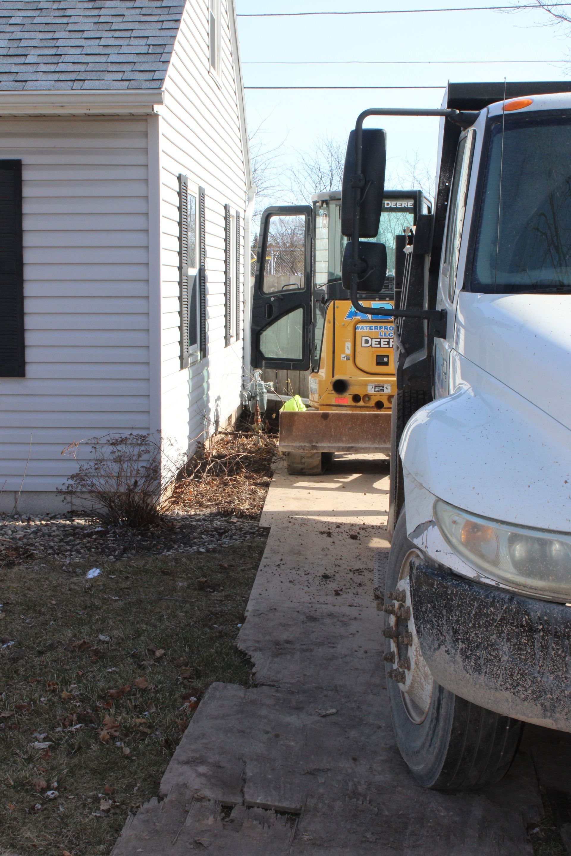 Yellow excavator and white truck parked next to a white house with a narrow walkway.