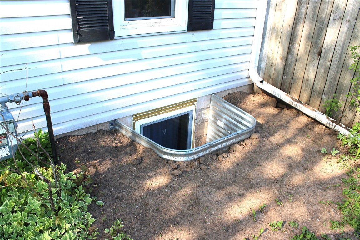 A corrugated metal window well at the base of a white house.