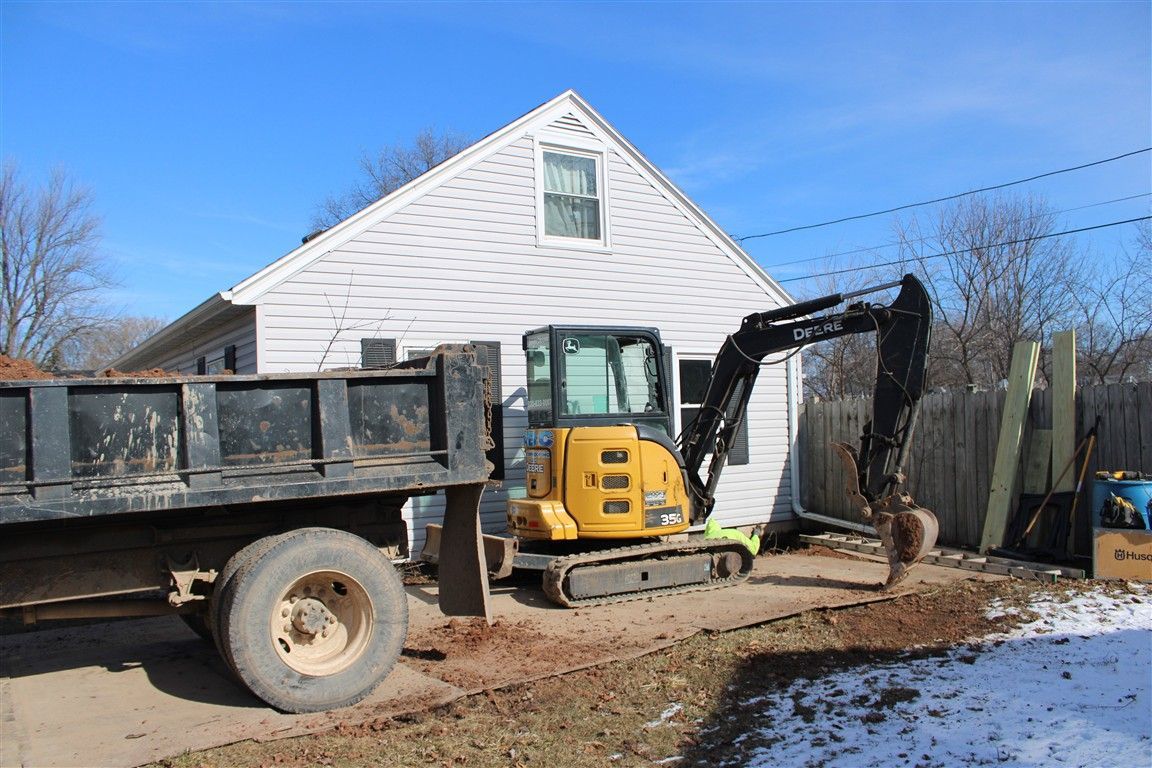 Yellow excavator digging near a house, next to a dump truck on a sunny day.
