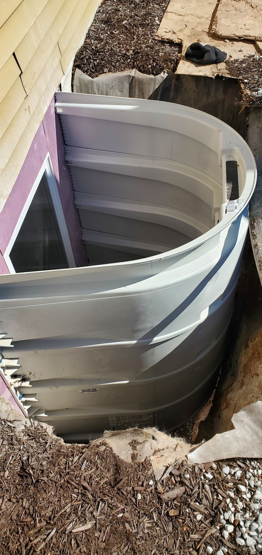 Curved gray window well. The well is in a dirt excavation next to a building.