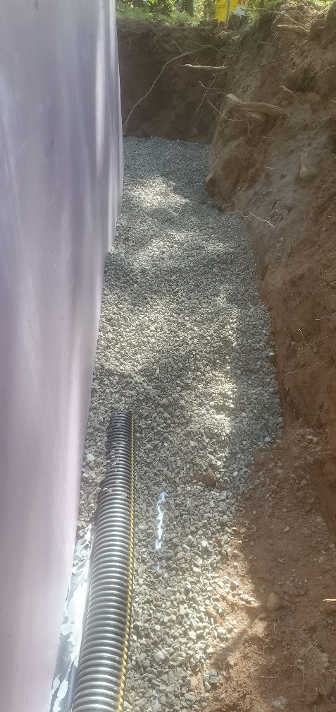 Trench filled with gravel next to a purple wall with a drainage pipe at the bottom.