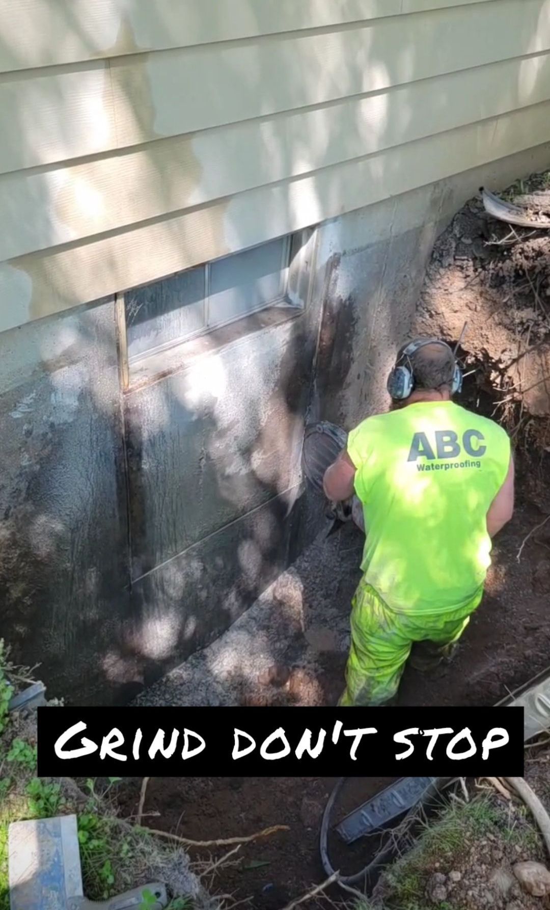 A worker in neon green gear using a grinder on a concrete foundation wall, outdoors.