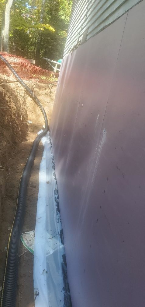 Exterior foundation waterproofing, drainage system being installed along a building wall, surrounded by dirt.