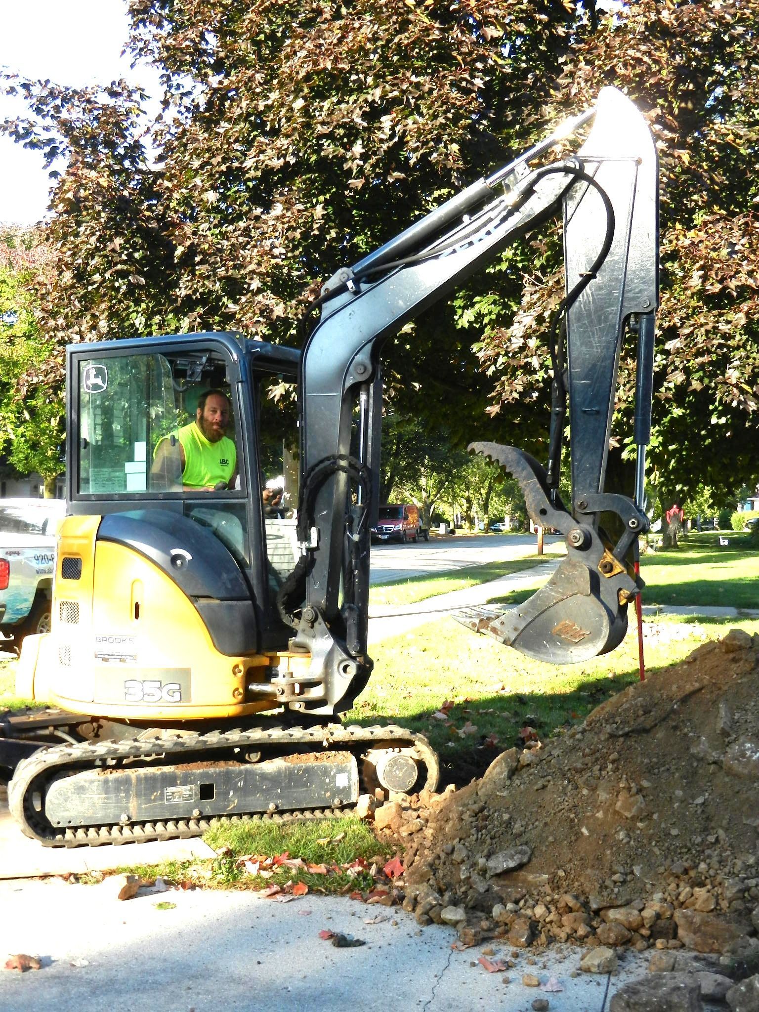 Yellow excavator digging near a street, an operator in the cab wearing a safety vest.