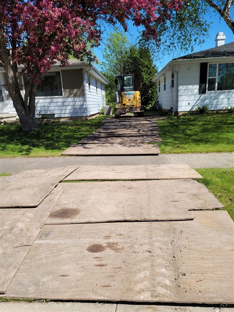 Plywood path leading to a yellow construction vehicle between two houses on a sunny day.