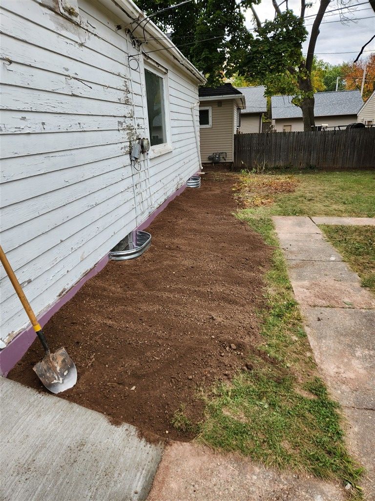 A newly tilled strip of dark soil next to a white house, sidewalk, and grass. 