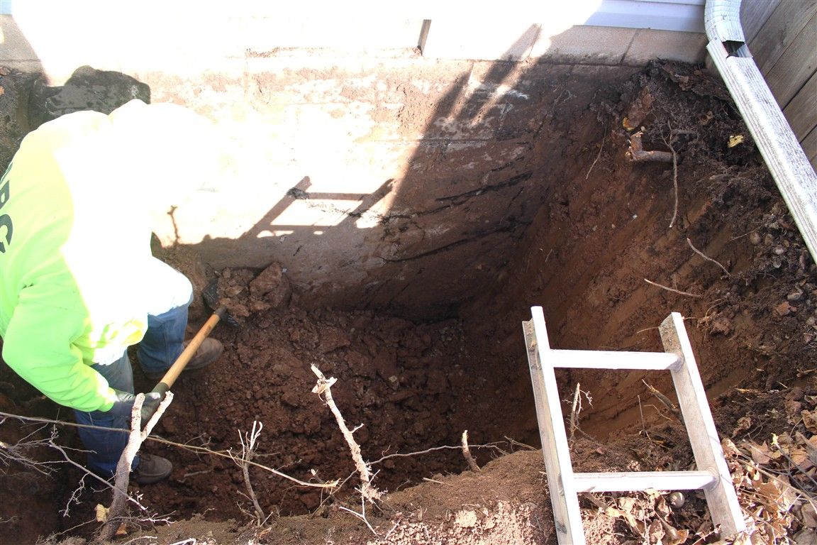 Person in yellow vest shoveling dirt in a deep rectangular hole next to a building. 