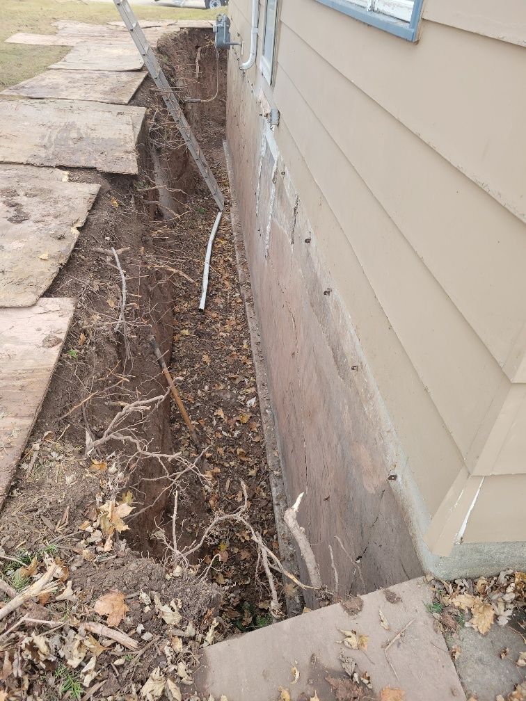 A trench dug along a building's beige siding, with wooden boards and a ladder.