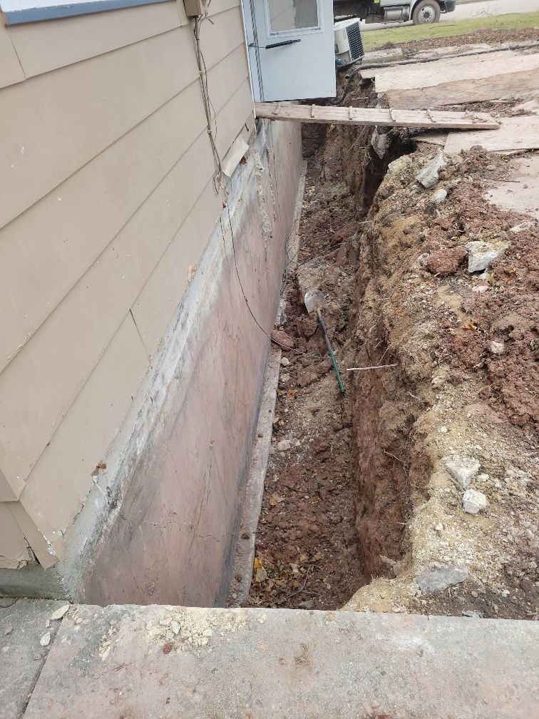 Excavation next to a house foundation, showing a deep trench.