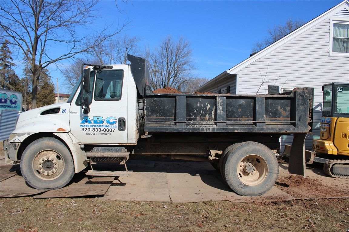 White dump truck parked on concrete, with dirt in the bed, next to a house and excavator.