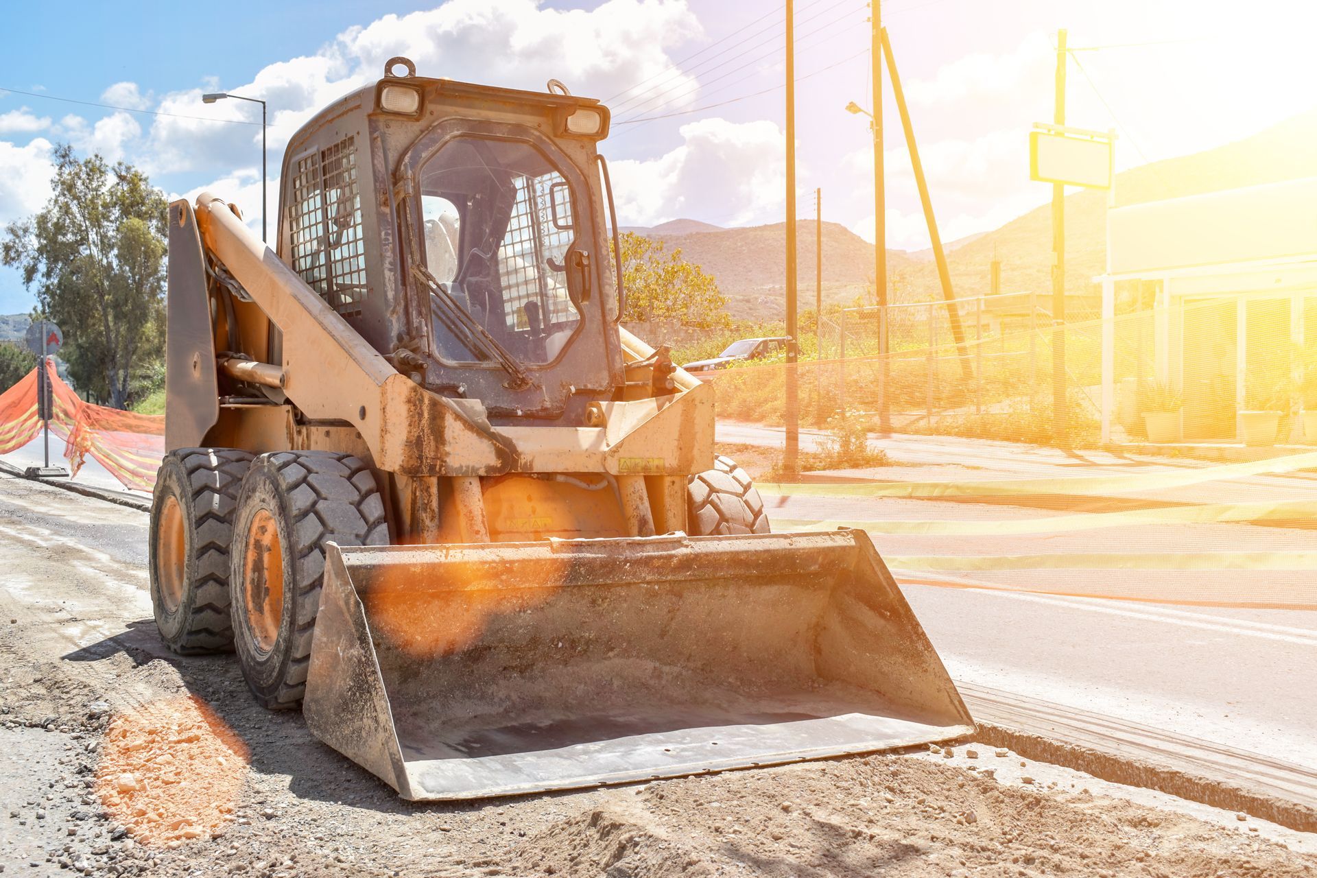 Yellow skid steer loader on a road construction site, scooping dirt in the sunlight.