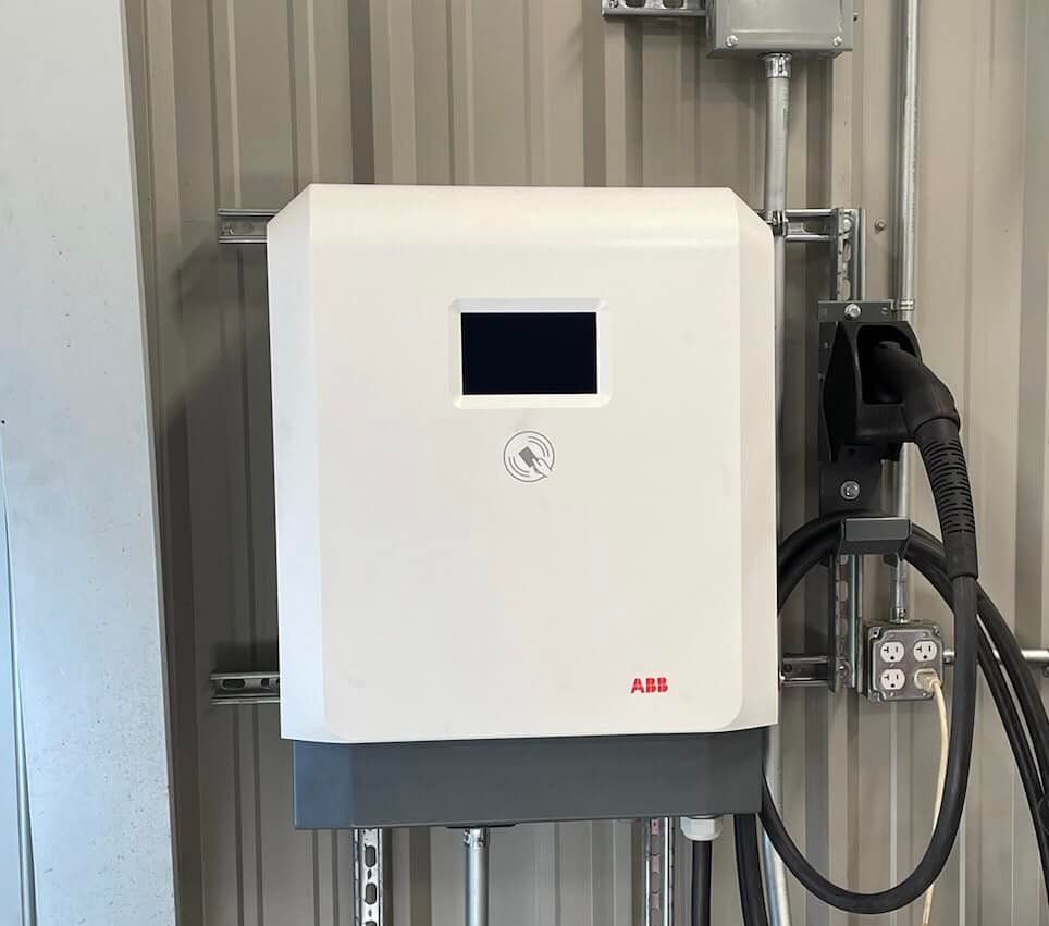 White ABB electric vehicle charger mounted on a metal wall with a charging cable and outlet.