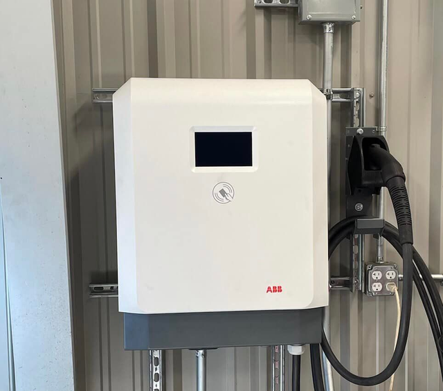 White ABB electric vehicle charger mounted on a metal wall with a charging cable and outlet.