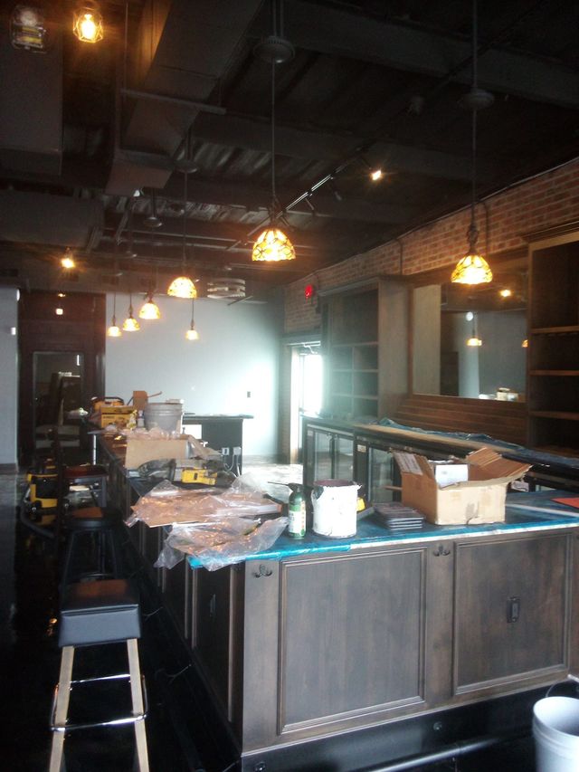 Interior of a bar under construction. Dark wood bar and shelving, tools scattered about, hanging lights.
