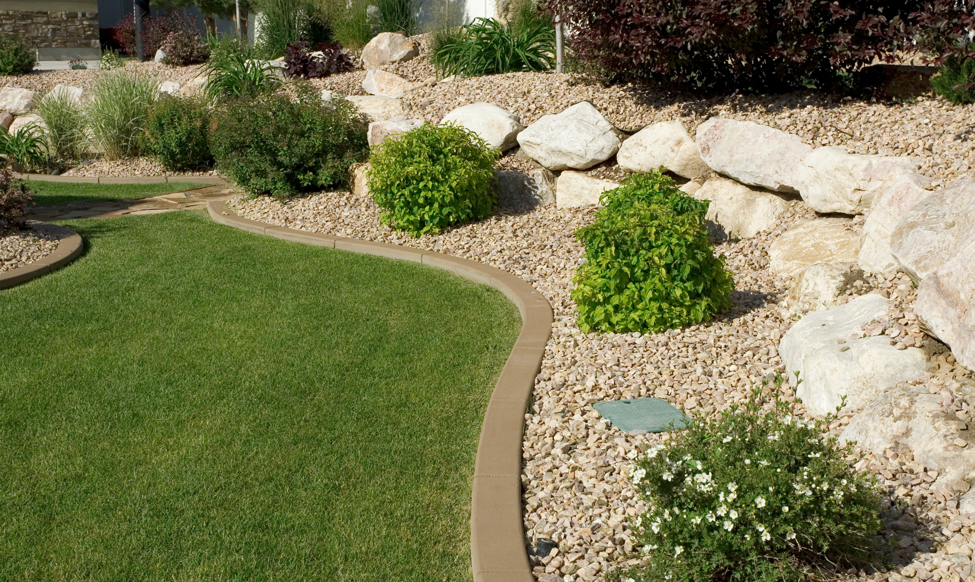 Lawn edged with a concrete border, adjacent to a rock garden and bushes.