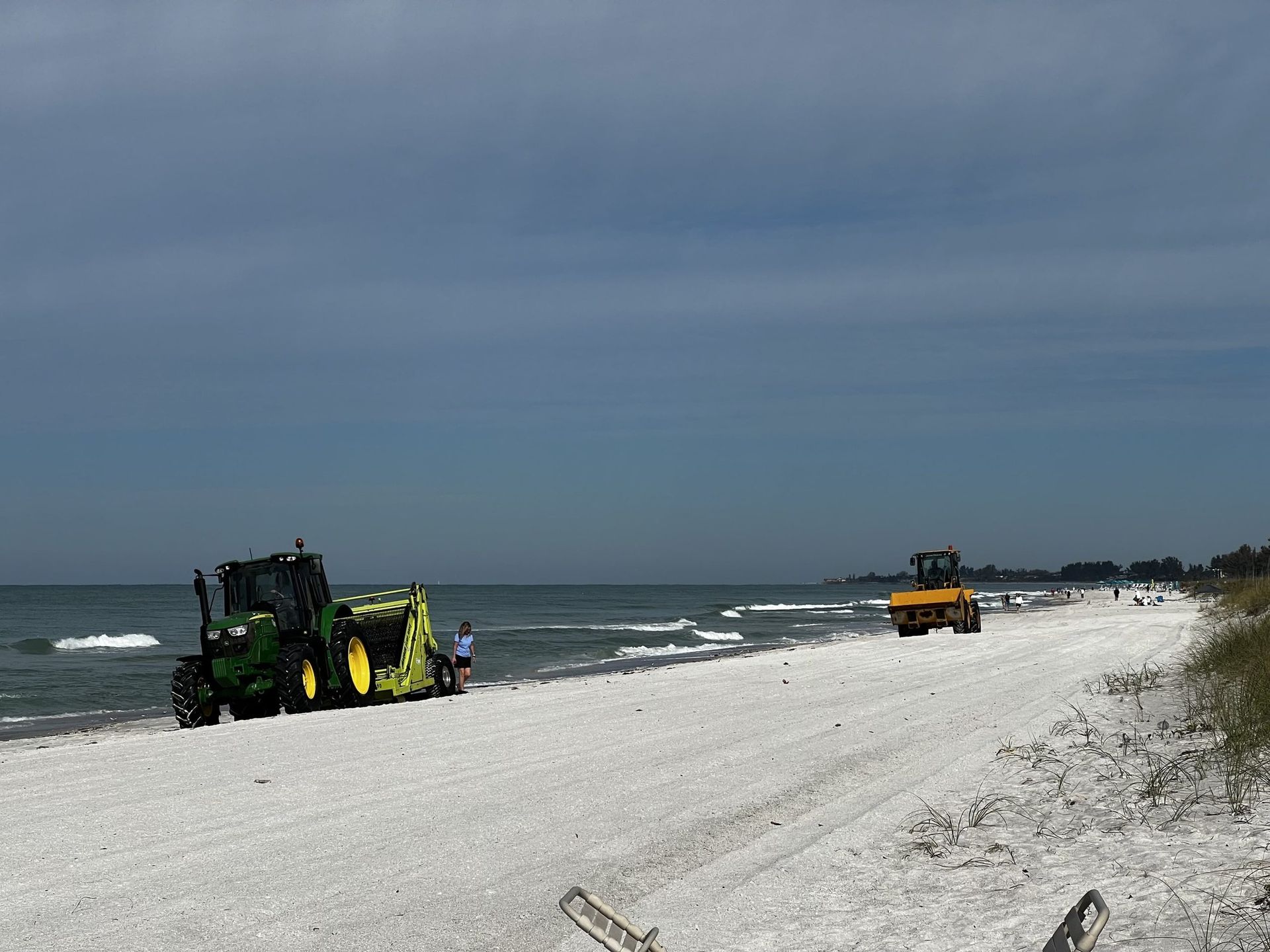 Two tractors on a sandy beach; one green, one yellow, by the ocean under a cloudy sky.