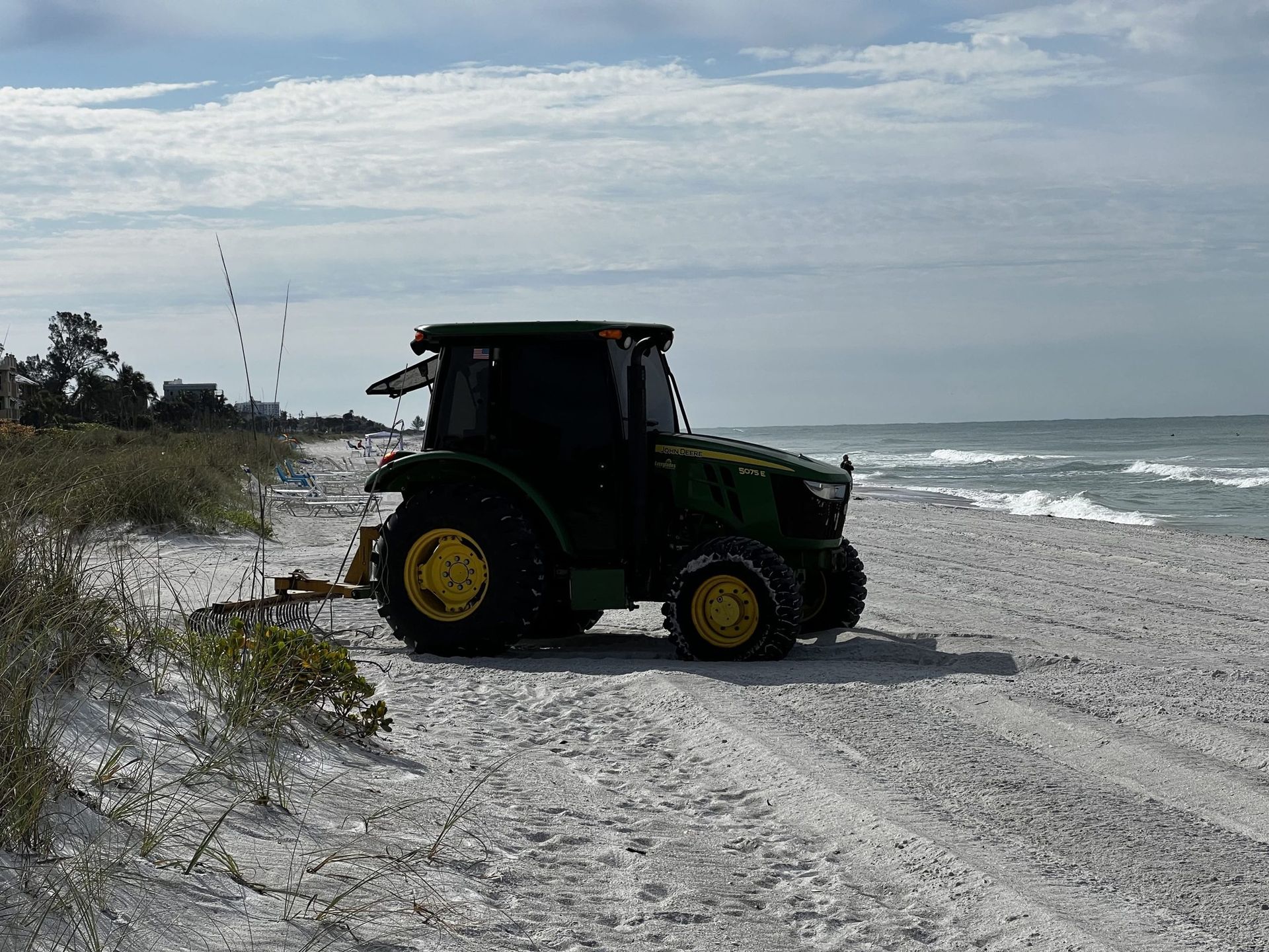 John Deere tractor on a sandy beach, ocean in the background, cloudy sky.