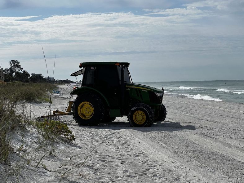 John Deere tractor on a sandy beach, ocean in the background, cloudy sky.