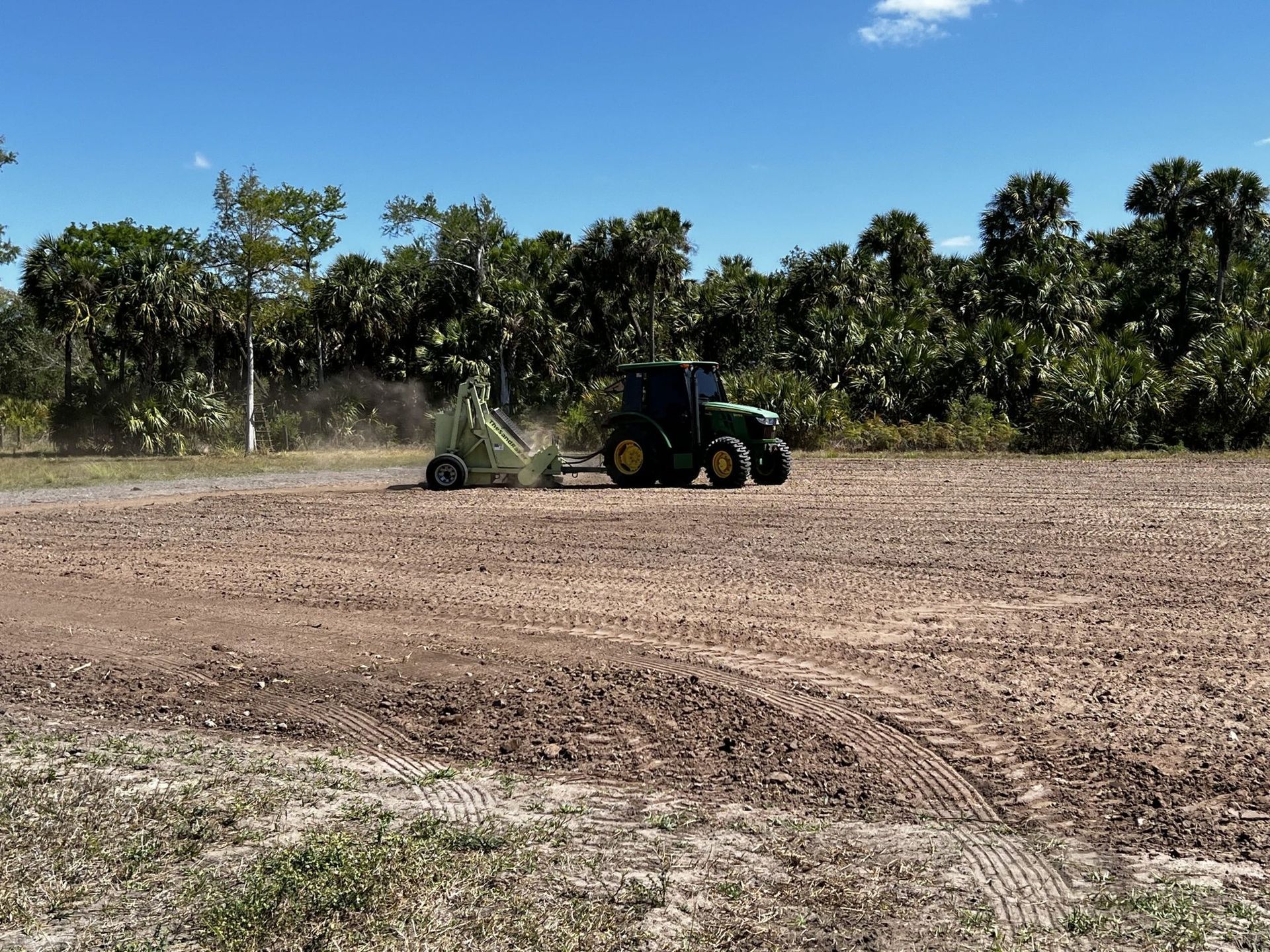 A green tractor rakes dirt in an open field under a blue sky with trees in the background.