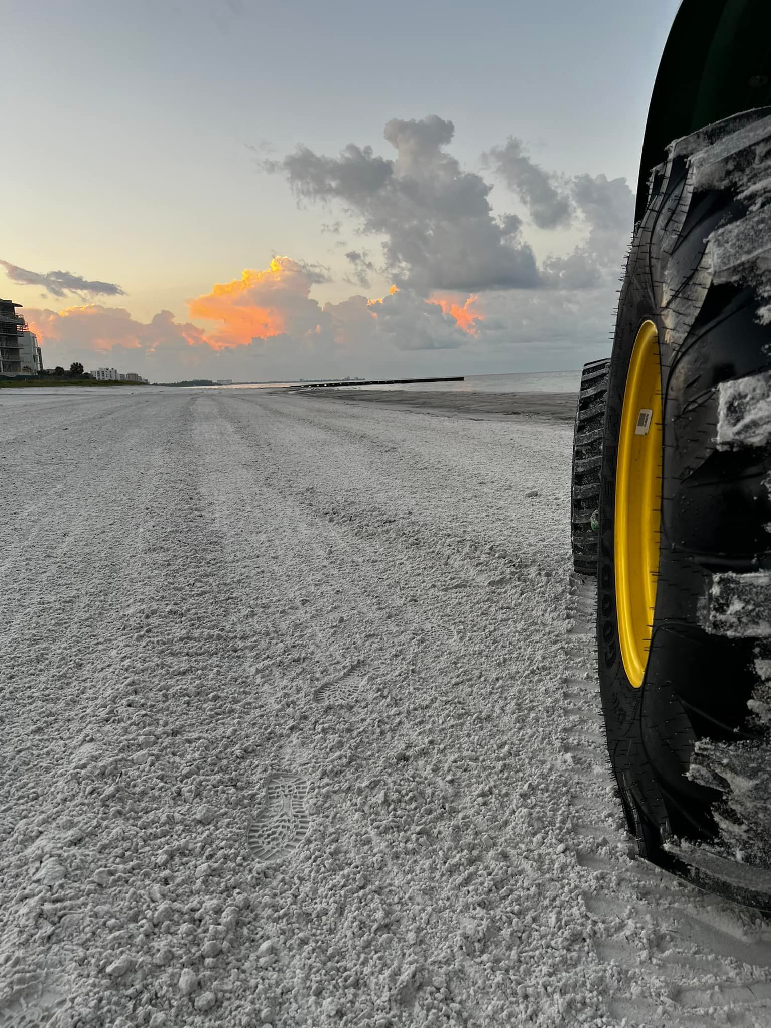 Tractor tire on a beach at sunset; yellow wheel, gray sand, pastel sky, ocean in the background.
