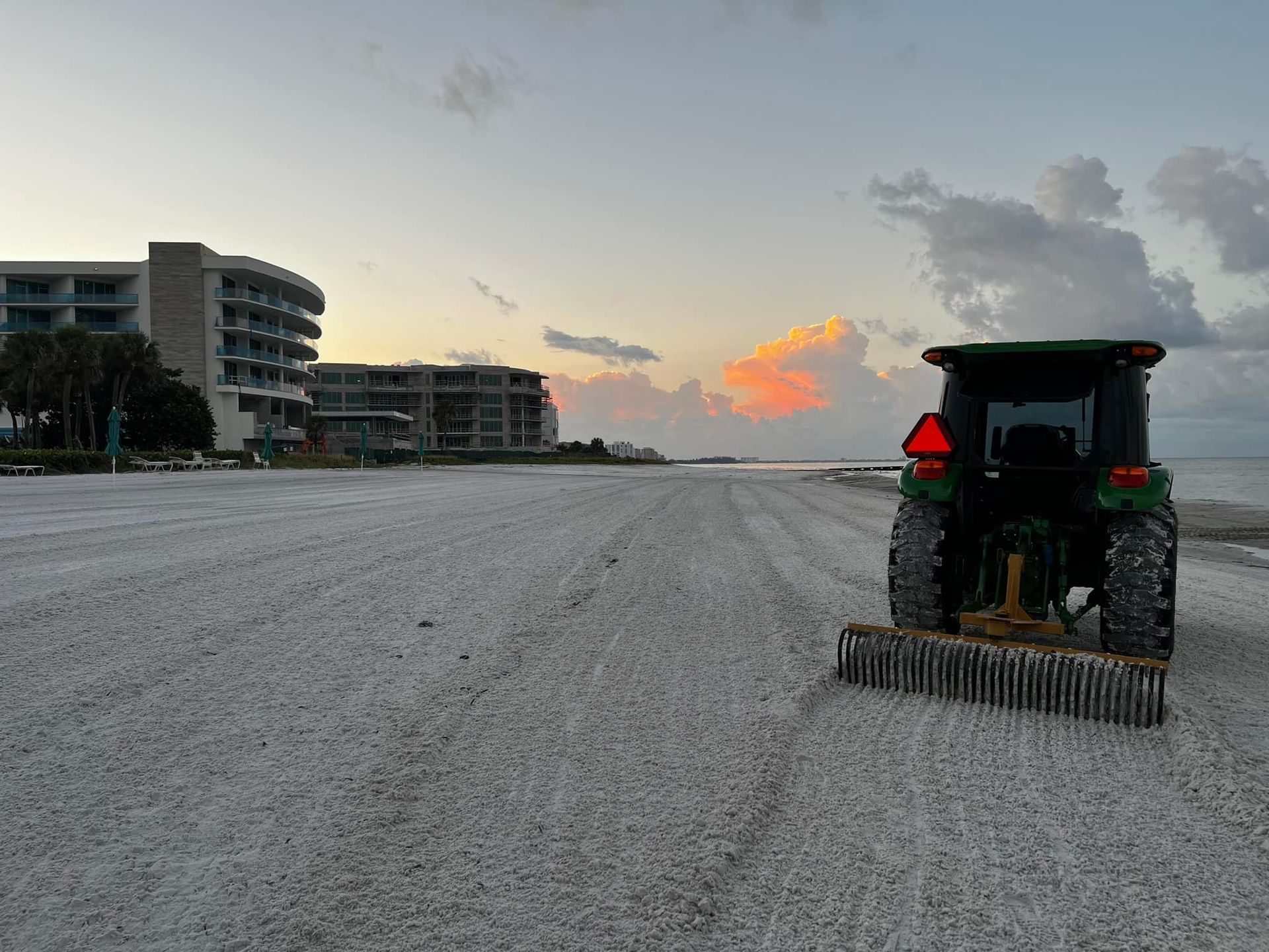 Tractor raking a beach at dawn, with a building in the background under a colorful sky.