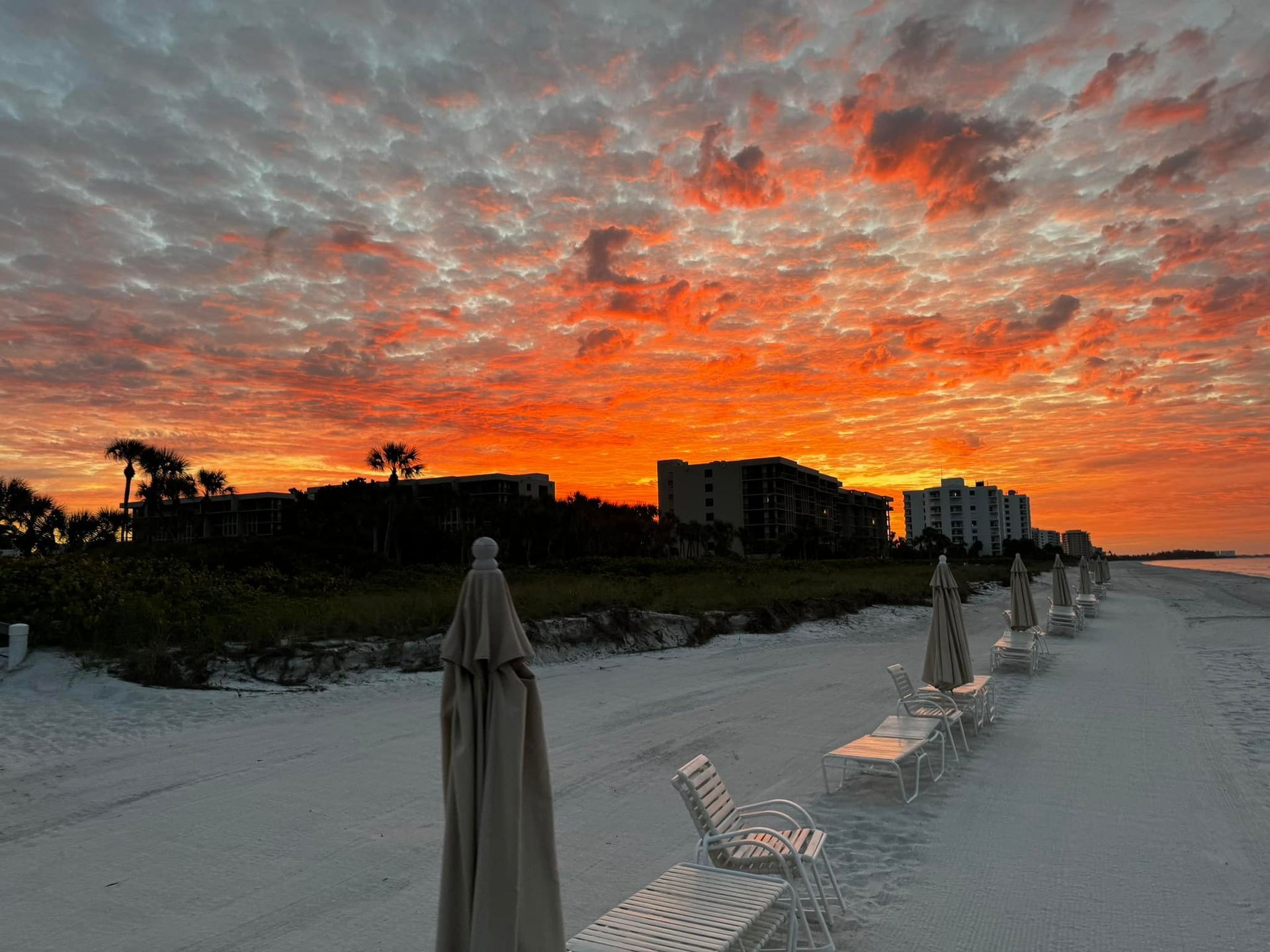 Sunset over beach with closed umbrellas and buildings silhouetted against orange and pink sky.