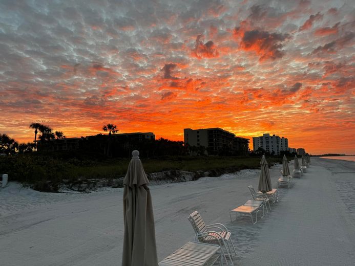 Sunset over beach with closed umbrellas and buildings silhouetted against orange and pink sky.