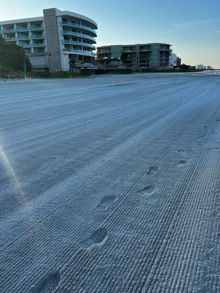 Beach with raked sand, footprints, and hotel buildings in the background. Blue sky, sunny day.