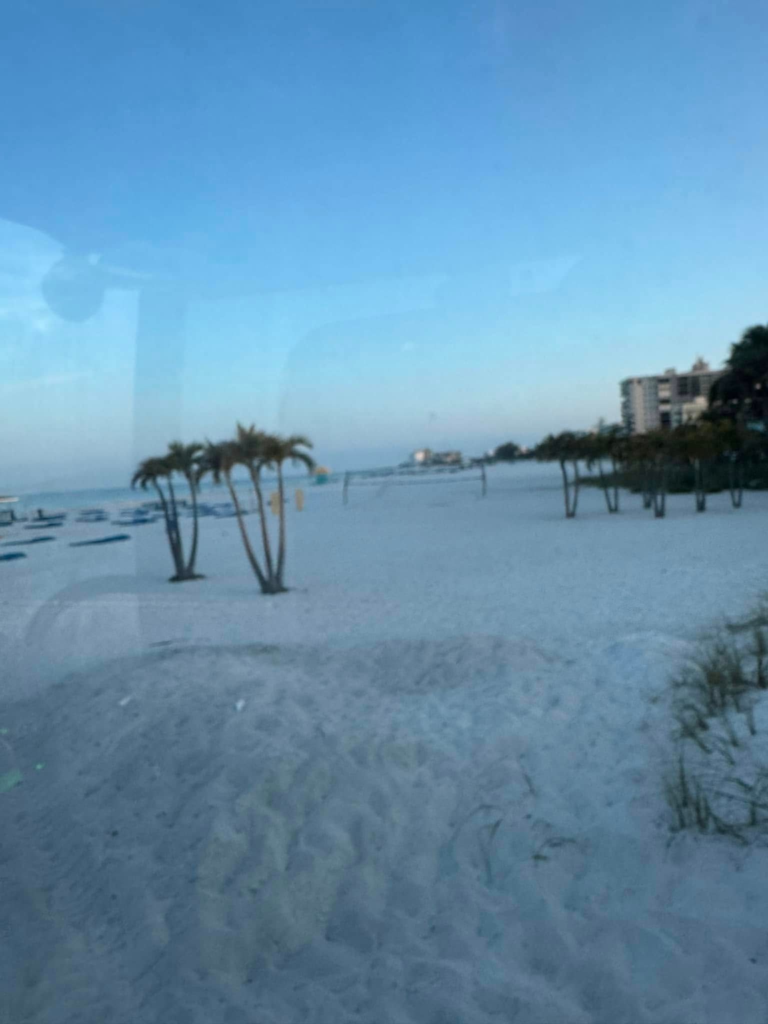 Beach with white sand, palm trees, and a clear blue sky. A building is in the distance.