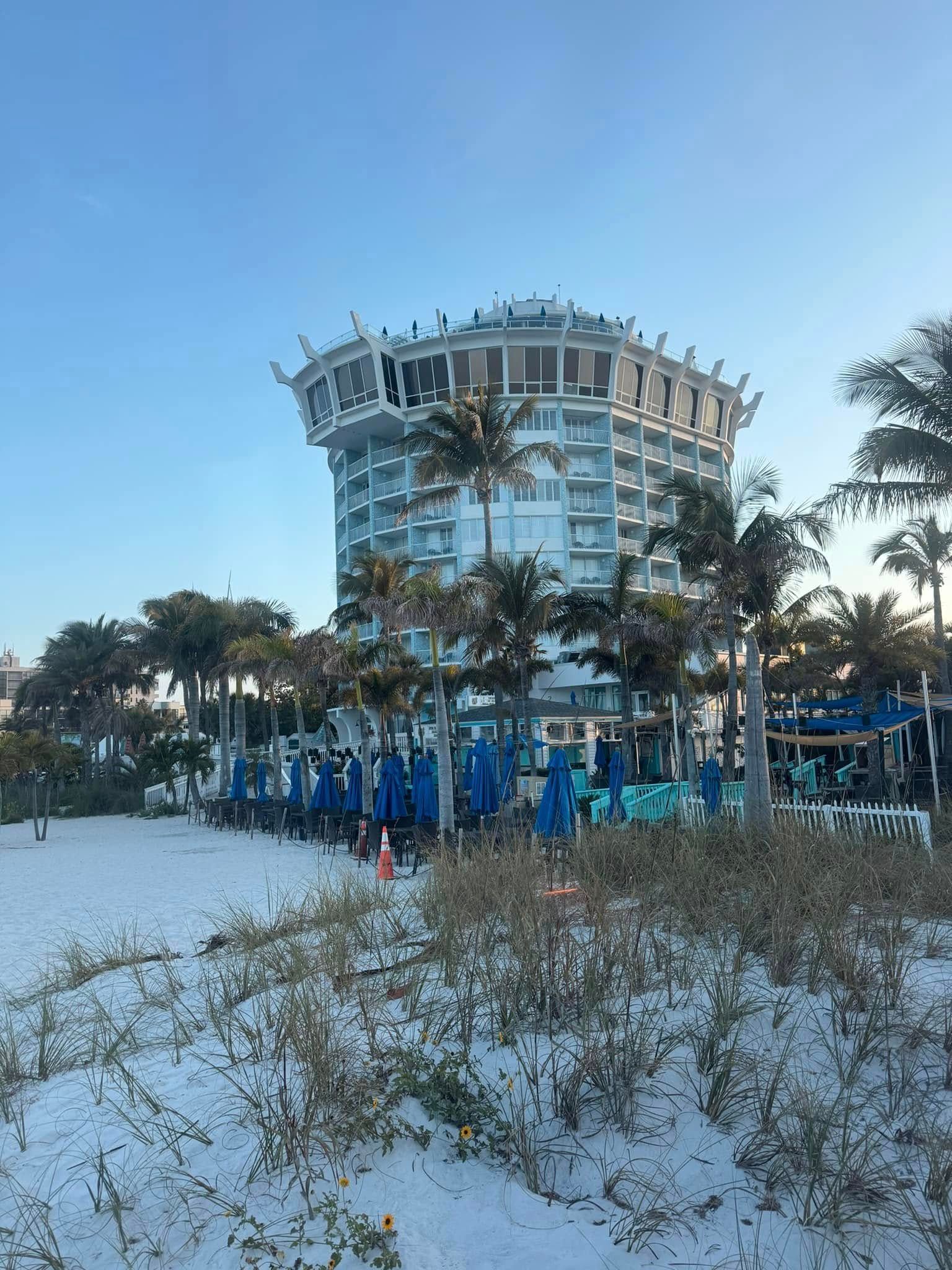 Beach scene with a tall, rounded building. Blue umbrellas and palm trees line the shore.