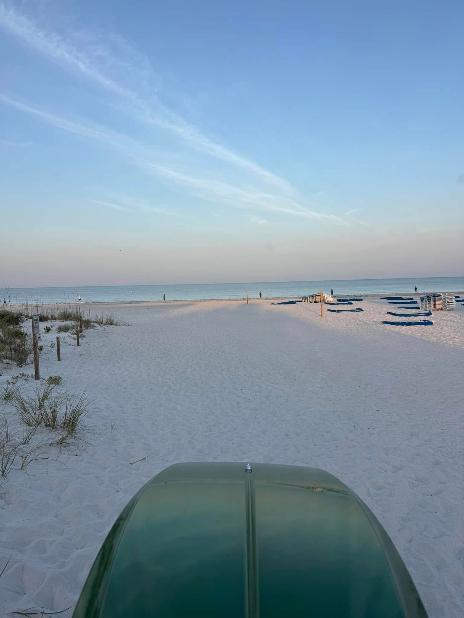 Beach scene with white sand, ocean, and blue sky; green car hood in foreground.