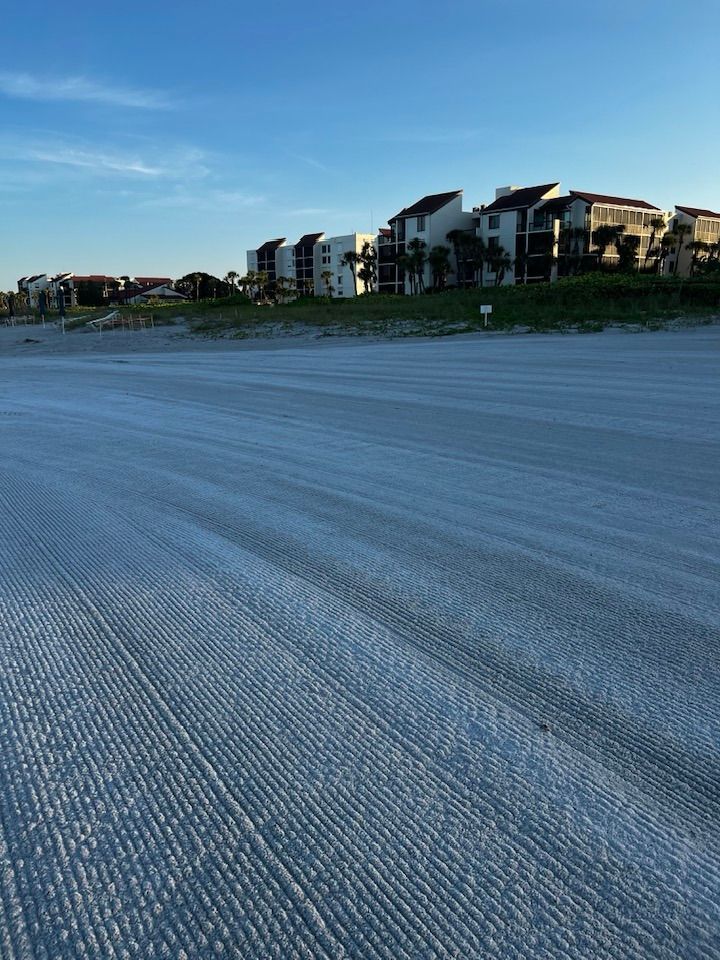 Sandy beach with tire tracks, buildings in the background, blue sky.