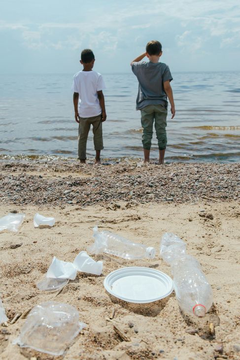 Two boys stand on a beach, looking out at the water; plastic trash litters the sand.