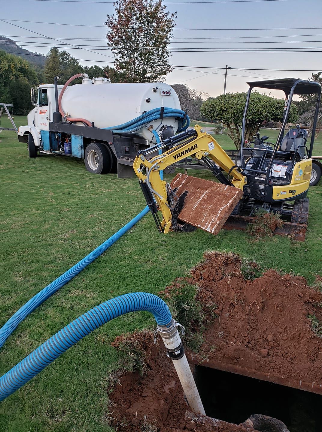 A tanker truck is pumping water into a hole next to a small excavator.