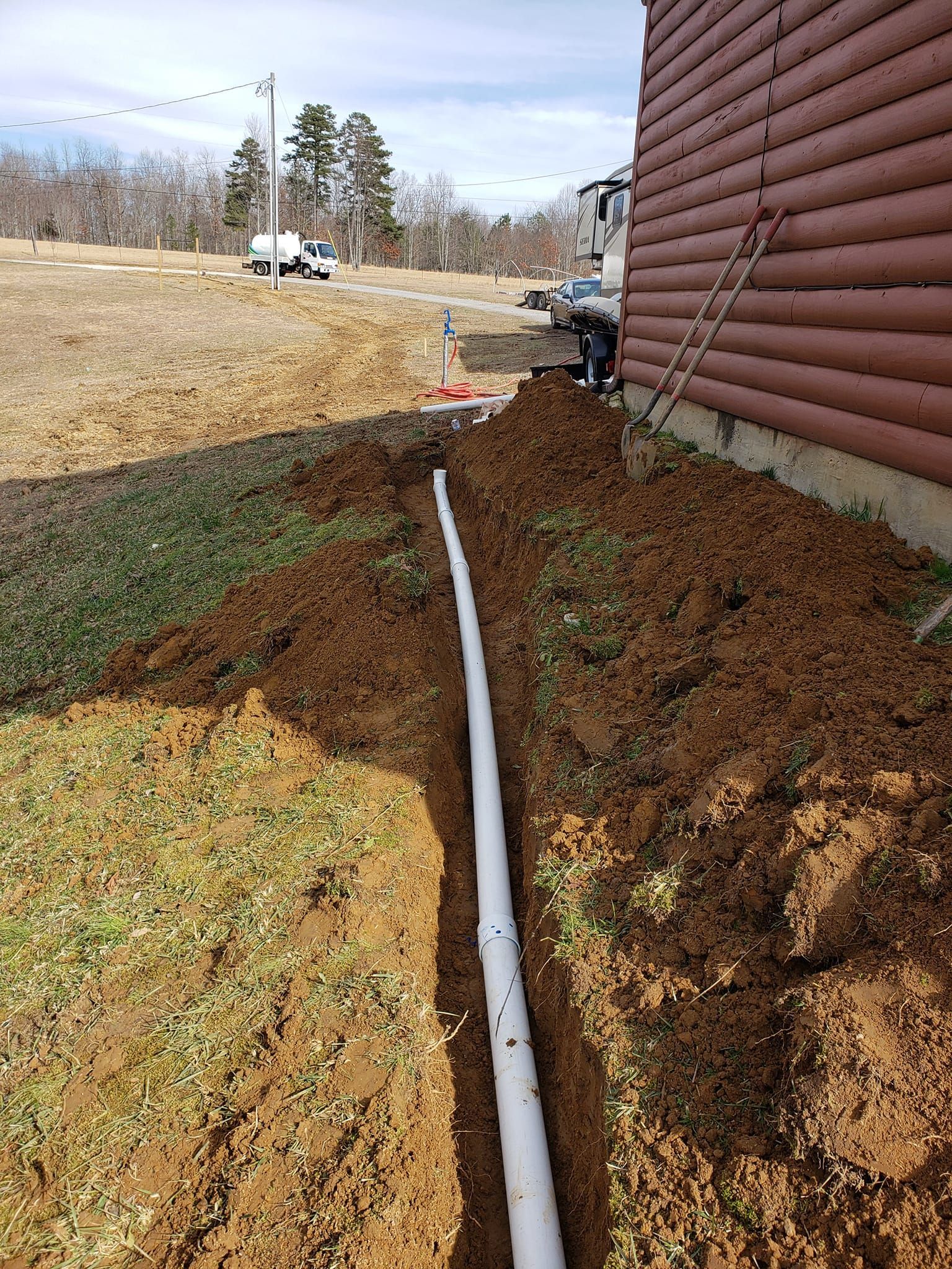 A white pipe is being installed in the dirt next to a log cabin.