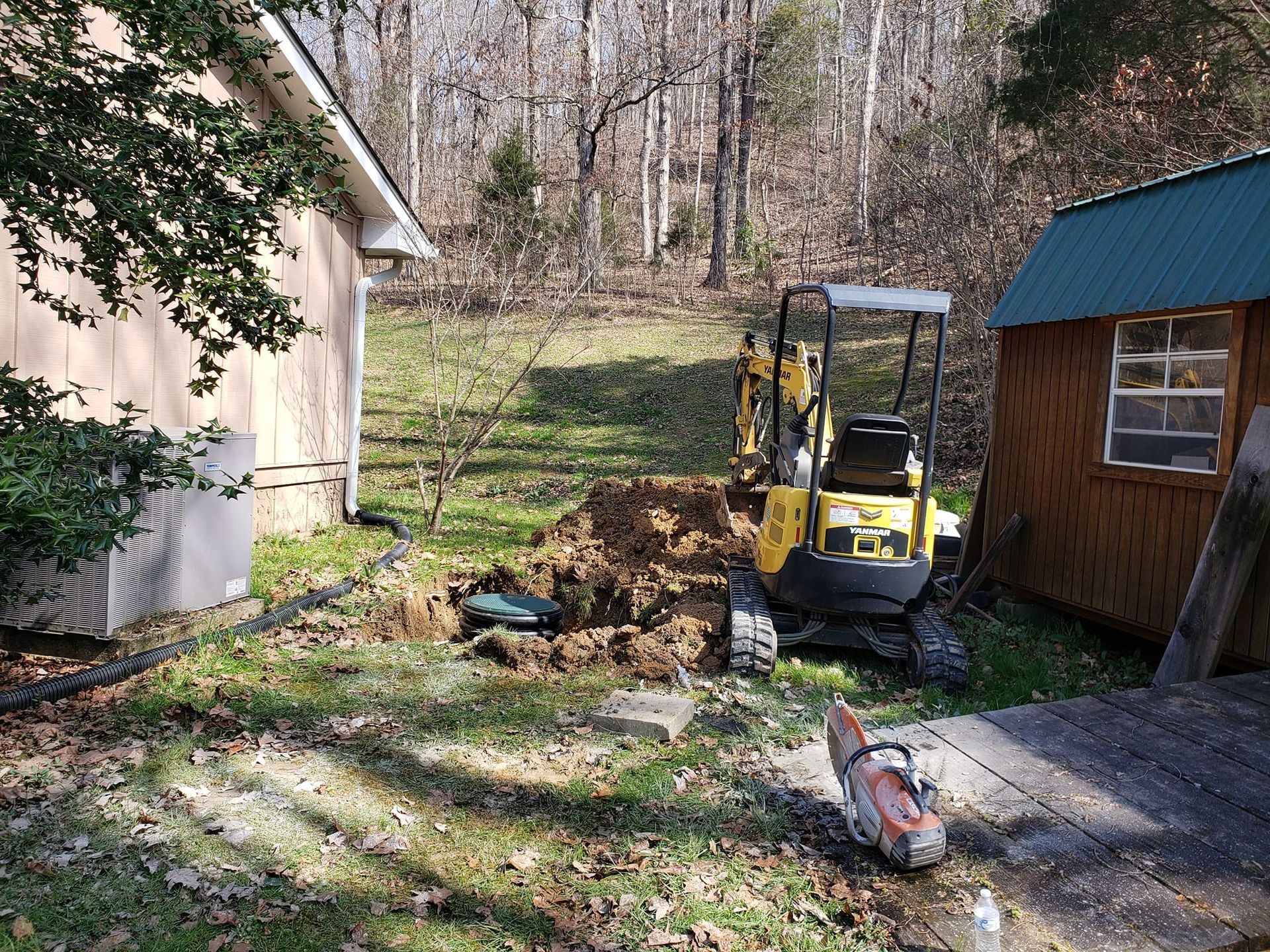 A yellow excavator is digging a hole in a yard next to a shed.
