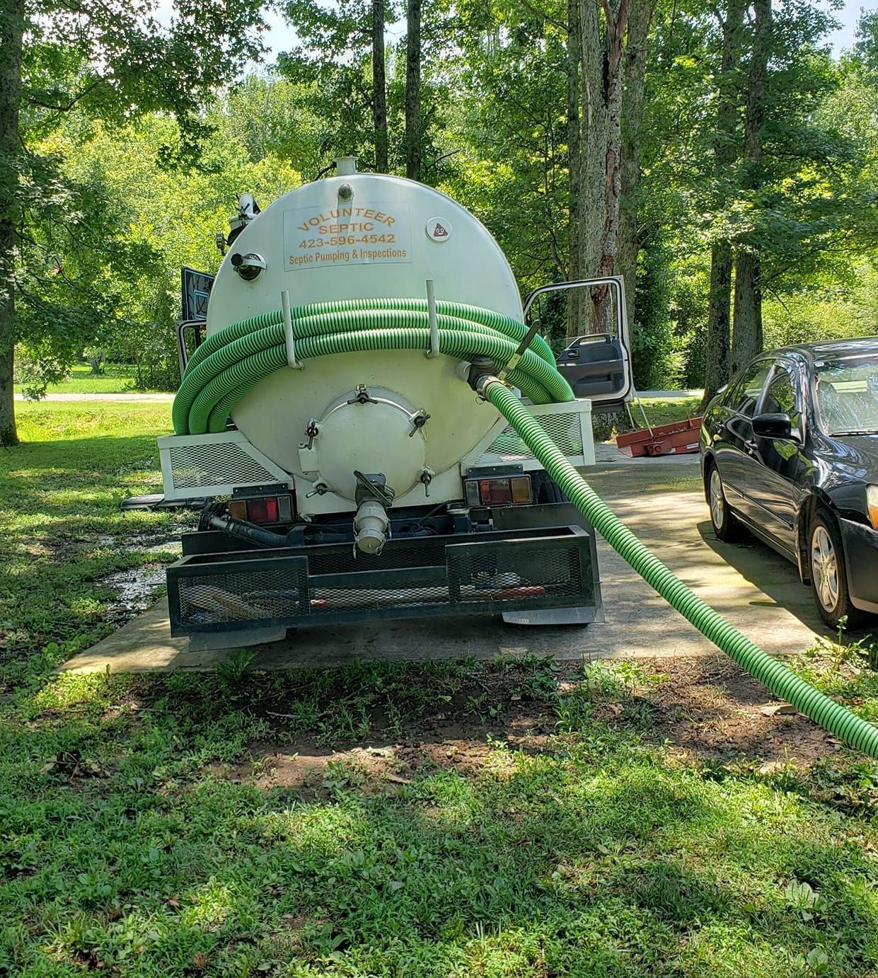 A septic tank truck with a green hose attached to it is parked in a driveway next to a car.