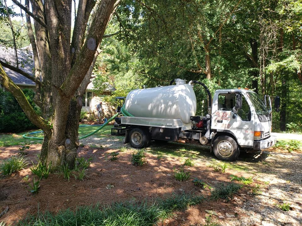 A white septic truck is parked in a driveway next to a tree.