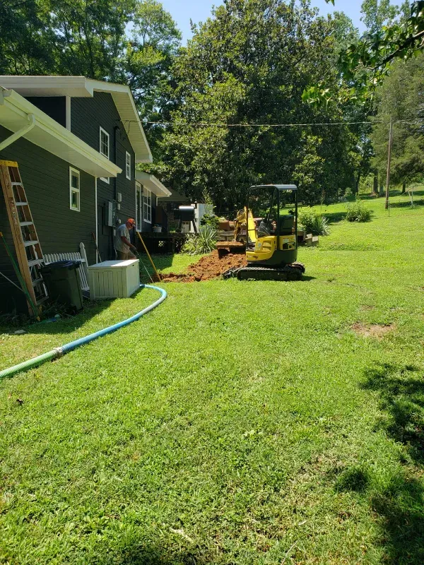 A yellow excavator is digging a hole in the grass in front of a house.