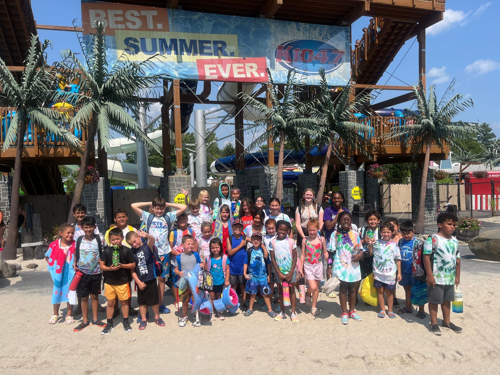 A group of children at a water park stand in front of water slides, under a sign that says 