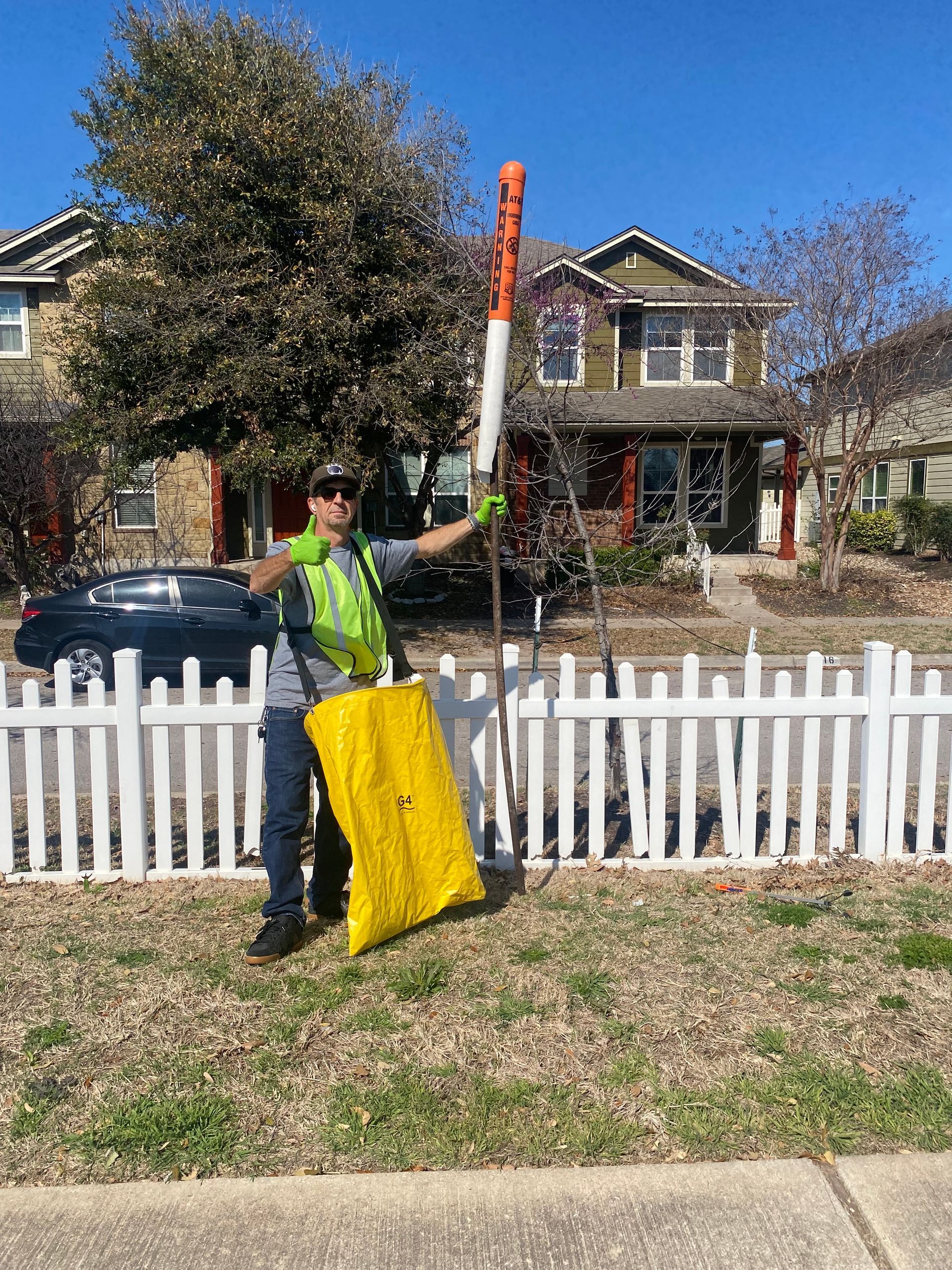 A man is standing in front of a white picket fence holding a shovel and a yellow bag.